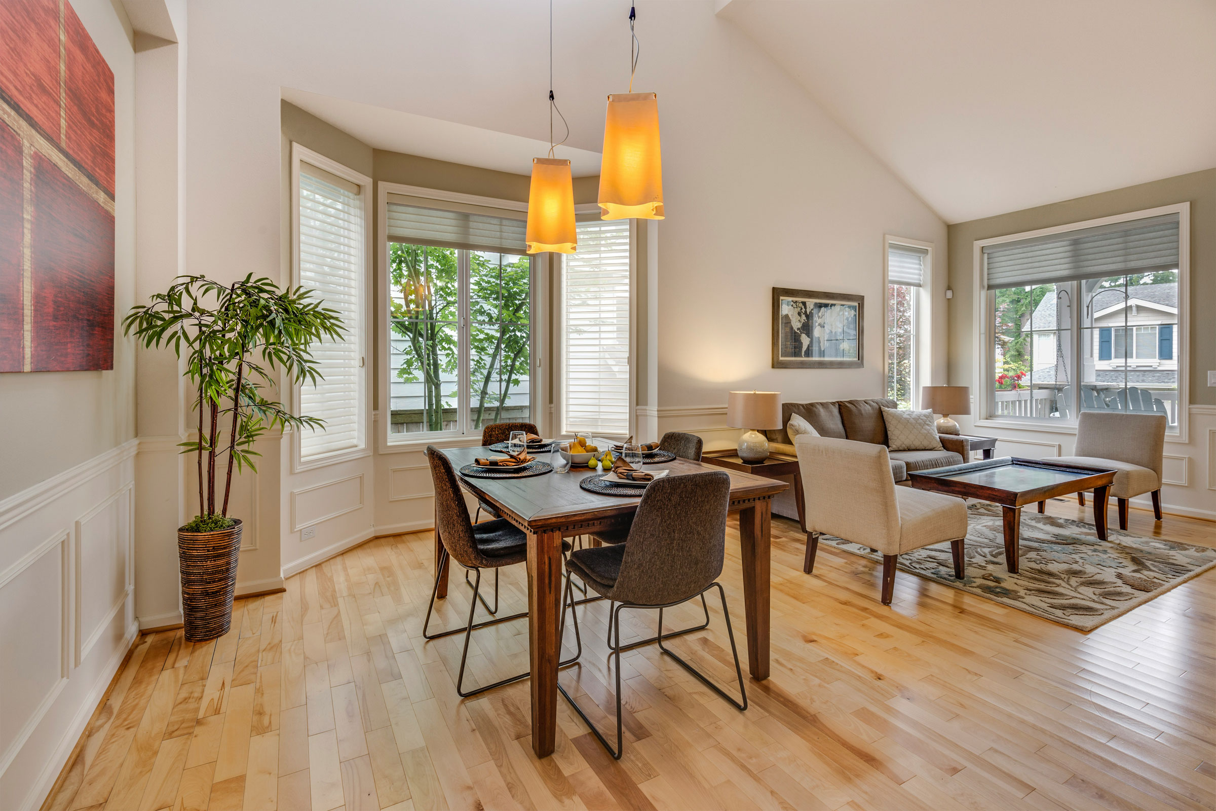 Bright open-concept dining and living room with vaulted ceilings, large windows, and light hardwood floors. Features a modern wood dining table with black and upholstered chairs, pendant lighting, a neutral-toned sectional sofa, and contemporary decor.