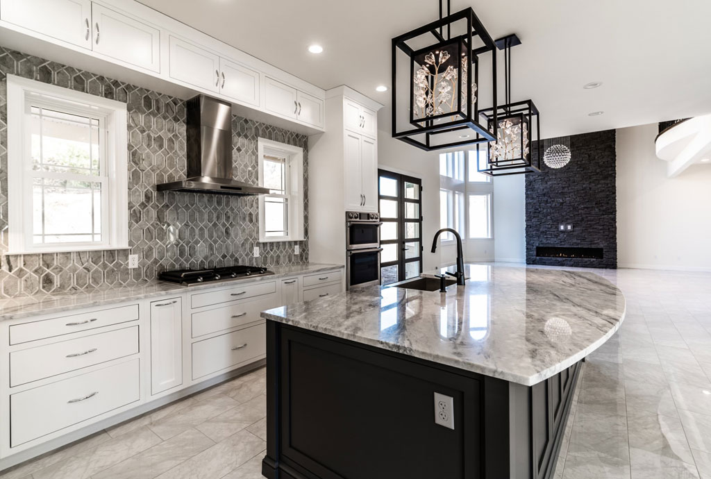 Striking modern kitchen featuring a large marble island with a curved edge and black cabinetry, complemented by dramatic geometric pendant lighting. The backdrop includes white shaker cabinets, a hexagonal gray tile backsplash, stainless steel range hood, and a sleek double oven. In the background, a contemporary black stone fireplace anchors the open-concept living space with high ceilings and abundant natural light.
