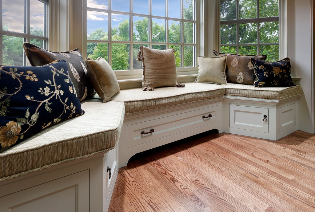 Cozy built-in window bench with cushioned seating and multiple throw pillows in earth tones and floral patterns. The bench features under-seat storage drawers and is framed by tall windows offering a scenic view of green trees and blue sky. Warm hardwood flooring completes the inviting nook.