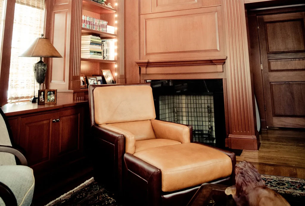 Cozy library or home office with rich wood paneling, built-in bookshelves, a leather armchair, and a fireplace with detailed molding.