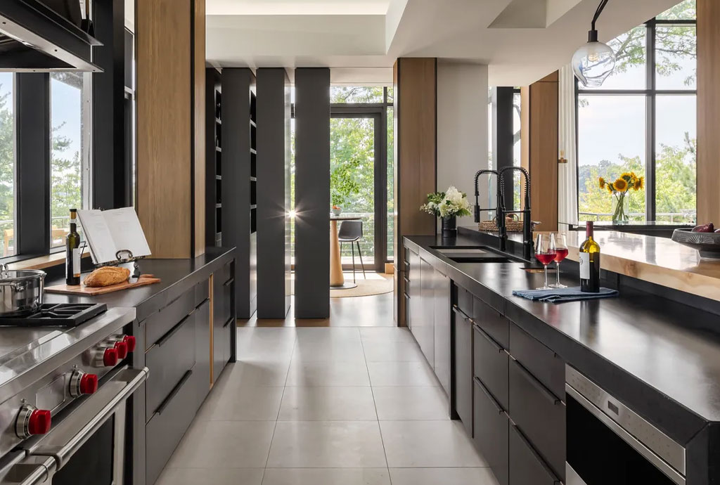 Modern open-concept kitchen with sleek black cabinetry, built-in appliances, a dual-sink island, and floor-to-ceiling windows overlooking lush greenery. A bottle of red wine, two glasses, and fresh flowers add a welcoming touch to the space.